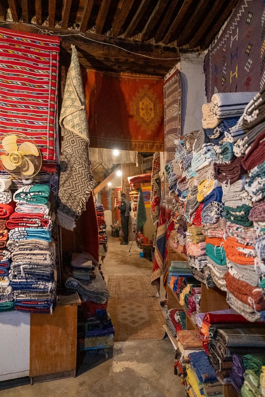 A carpet merchant at the back of his shop between walls of stacked kilims and folded Berber blankets, Fes medina Morocco