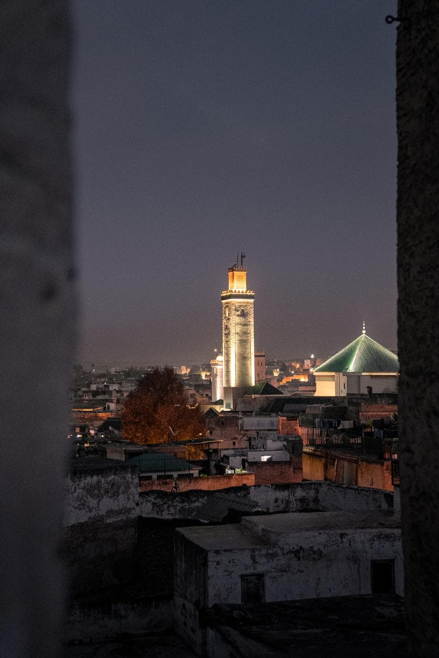 The lit minaret and green pyramid roof of the Kairaouine mosque above the rooftops of Fes el-Bali at dusk, Morocco