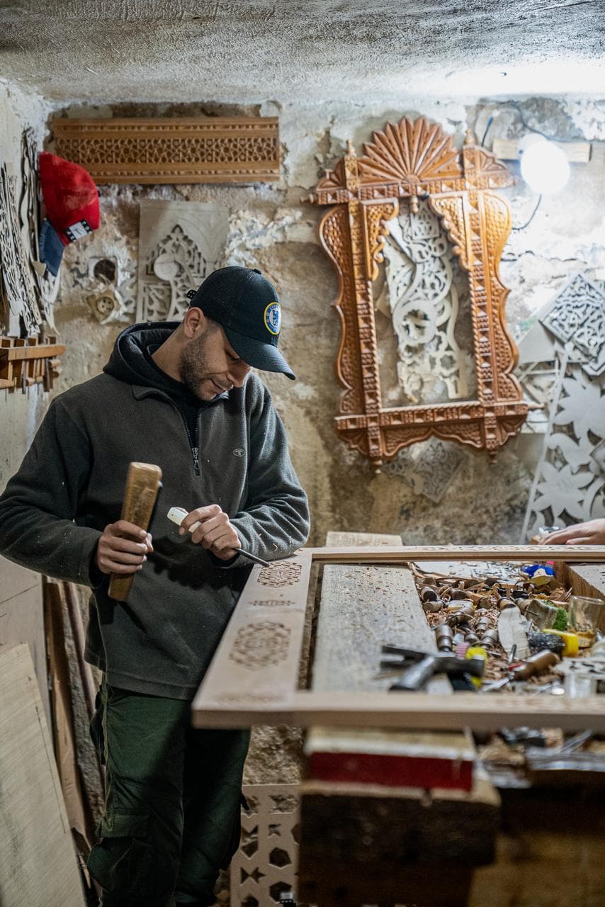 A woodcarver cutting a geometric rosette into a cedar panel with chisel and mallet in his workshop, Fes medina Morocco