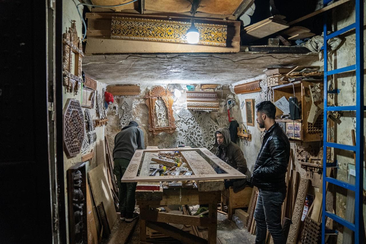 Three craftsmen at work on a carved cedar panel in a woodcarving workshop in the Fes medina, finished mashrabiya screens covering the walls, Morocco