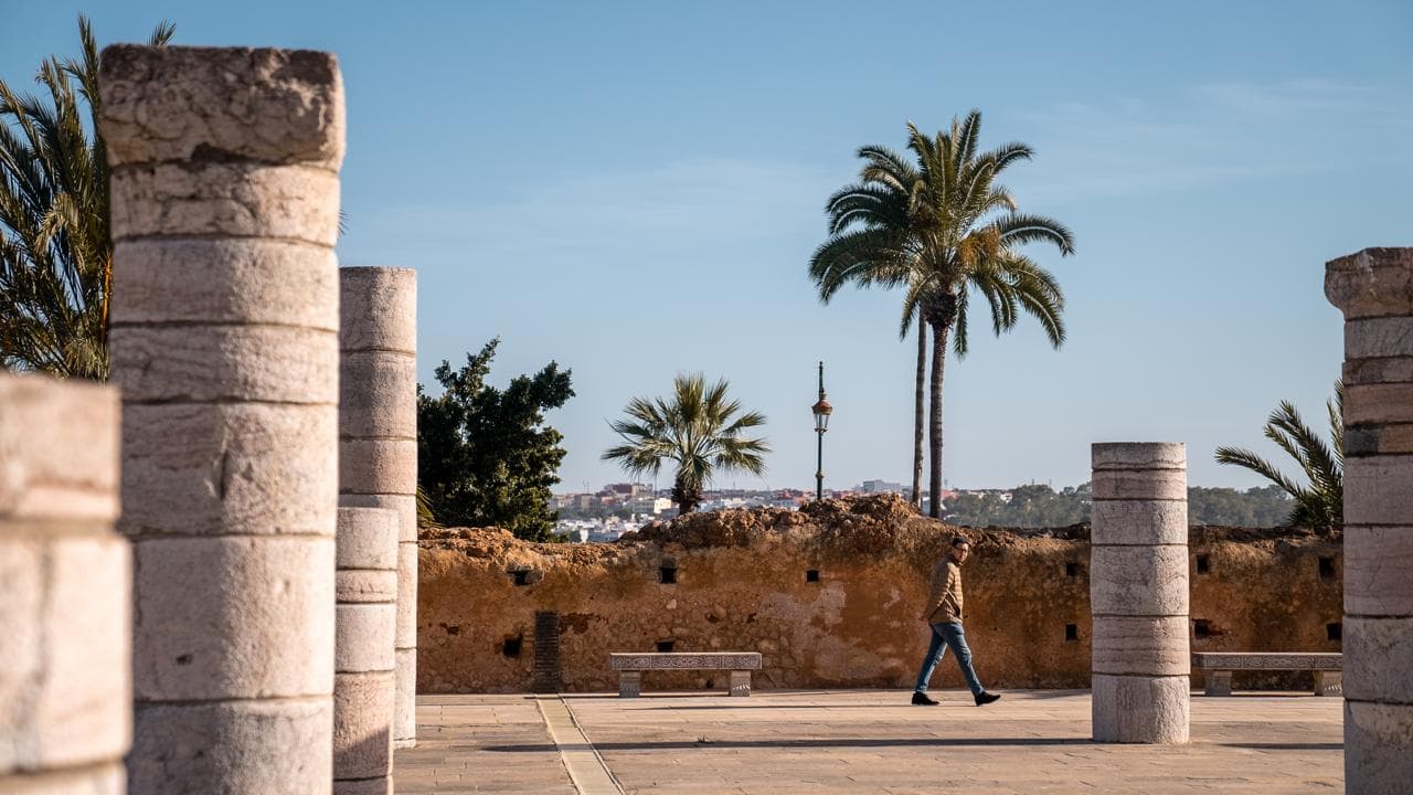 A visitor walking between the ranks of stone column stumps on the Hassan esplanade, with the Kasbah wall and Rabat beyond, Morocco