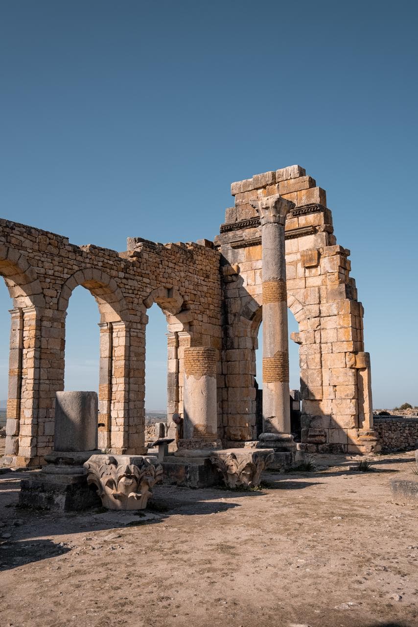 Fallen Corinthian capitals in the foreground of the standing basilica arcade at Volubilis, Morocco